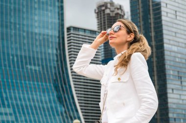 Beyaz ceketli ve güneş gözlüklü gülümseyen sarışın bir kadının görüntüsü.