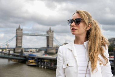 Beyaz ceketli ve güneş gözlüklü gülümseyen sarışın bir kadın görülüyor. Arka planda Thames ve Tower Bridge var.