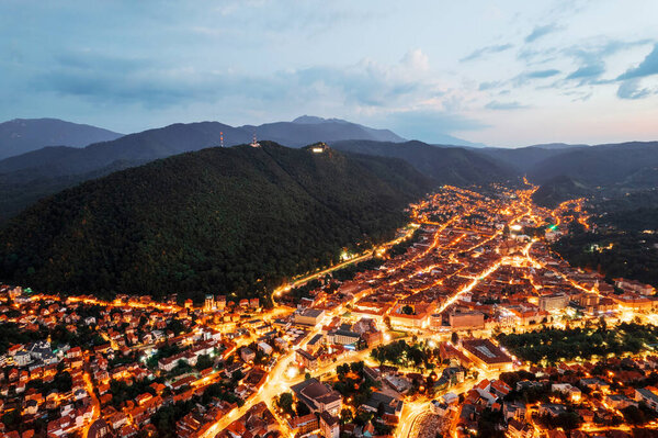 Aerial drone view of Brasov at sunset, Romania. Old city centre with old buildings and illumination, hills covered with greenery