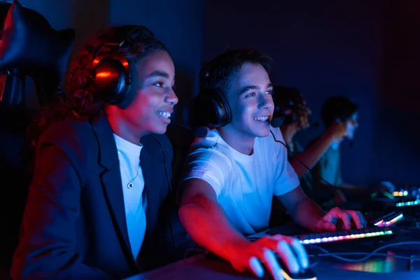 White boy and black girl teens in headsets playing video games in video game club with blue and red illumination. Keyboard and mouse with illumination