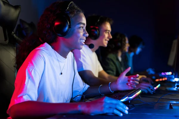 Multiracial group of teens in headsets playing video games in video game club with blue and red illumination. Keyboards and mice with illumination