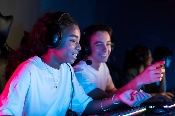 White boy and black girl teens in headsets playing video games in video game club with blue and red illumination. Keyboard with illumination