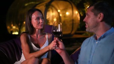 Şarap içen ve geceleri şöminenin yanında konuşan mutlu bir çift. Glamping arka planında şeffaf kabarcık çadırı