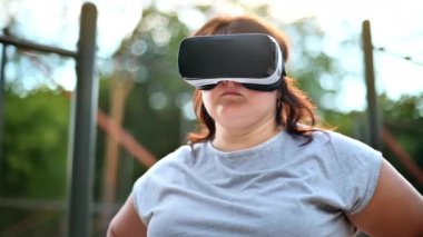 Kilolu bir kadın parkta spor sahasında sanal gerçeklik kulaklığı takıyor.