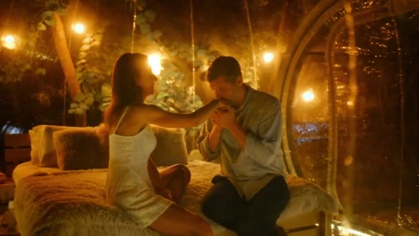 一对夫妇坐在透明的泡沫帐篷里 夜晚的釉面上有灯光 男人亲吻他妻子的手 互相拥抱 背景上的茂密森林 — 图库视频影像
