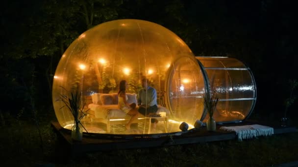 一对夫妻坐在透明的泡泡帐篷里聊天 晚上在玻璃窗上点亮灯光 背景上的茂密森林 — 图库视频影像