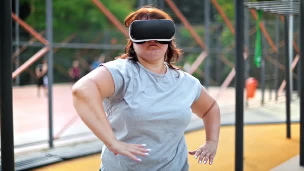 仮想現実のヘッドセットを着用しながら 公園のスポーツフィールドで運動する灰色のTシャツとレギンスの太りすぎの女性 フィットネスライフスタイル — ストック動画