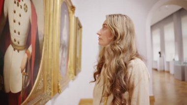 CHISINAU, MOLDOVA - 21 Eylül 2023: Bir sanat galerisinde yürüyen ve duvarlardaki birden fazla tabloya bakan sarışın bir kadın. Yavaş çekim