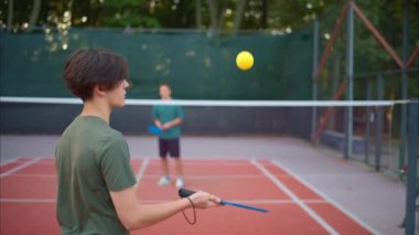 Sahada sarı top ve mavi raket oynayan çocuklar.