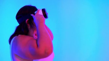 Eşofmanlı aşırı kilolu bir kadın ve mavi arka planı ve kırmızı ışığı olan bir stüdyoda VR kulaklıklı bir kadın.