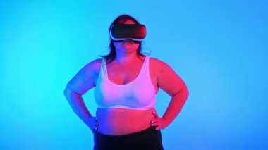 Aşırı kilolu eşofmanlı ve VR kulaklıklı bir kadın stüdyoda poz veriyor mavi arka plan ve kırmızı ışık, kameraya bakıyor.