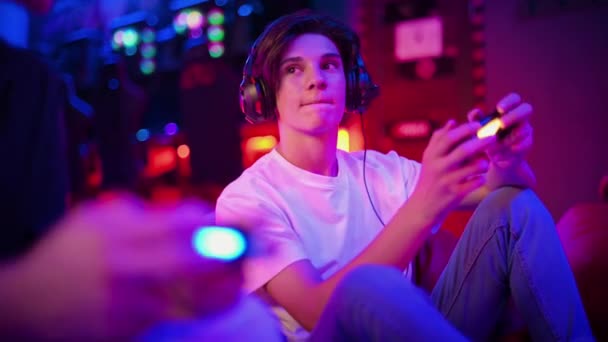 ヘッドフォンにいる2人の10代の友人が ビーンバッグに座ってゲームコンソールを使って話したり 話したり 笑ったりしている 青と赤のイルミネーション — ストック動画