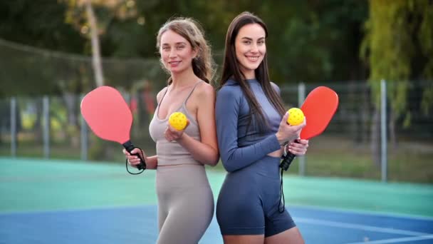 两名身穿运动服 面带微笑的妇女在室外场地上打皮球时 摆出一副球和球拍的姿势 慢动作 — 图库视频影像