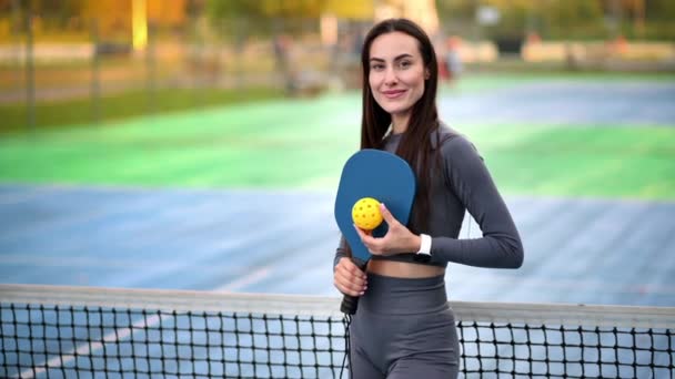 屋外コートでピックルボールをプレーするためのボールとラケットをポーズするスポーツスーツの笑顔の女性 スローモーション — ストック動画