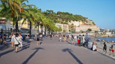 Nice, Fransa - 25 Eylül 2023: Birden fazla yürüyen insan ve yeşillikle gezinti, Akdeniz kıyısında dinlenen insanlarla plaj, arka planda klasik binalar