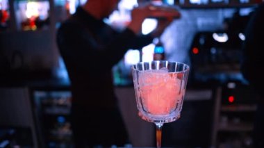 Barmen sallanıyor ve turuncu kırmızı alkollü kokteyl döküyor. Geceleri neon ışıkları olan bir barda buzlu.