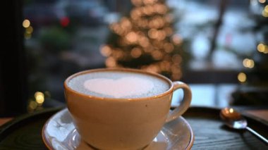 Kalp şeklinde köpüklü latte sanatı olan bir fincan kahve. Arka planda Noel ağacı