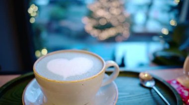Kalp şeklinde köpüklü latte sanatı olan bir fincan kahve. Arka planda Noel ağacı