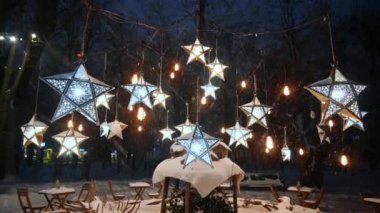 Kar yağarken yıldız şeklindeki fenerlerin ve parktaki kahve masalarının geniş açılı görüntüsü. Noel süsleri. Kış akşamı