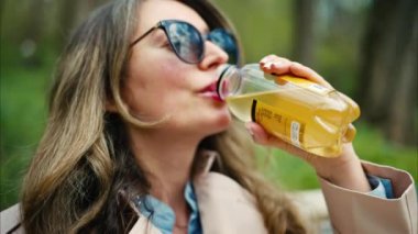 Esmer, genç bir kadın parkta bankta çay içiyor.