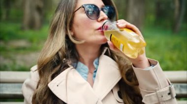 Esmer, genç bir kadın parkta bankta çay içiyor.