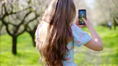 Mavi elbiseli esmer kadın, çiçek açan badem ağaçlarının arasında fotoğraf çekiyor.