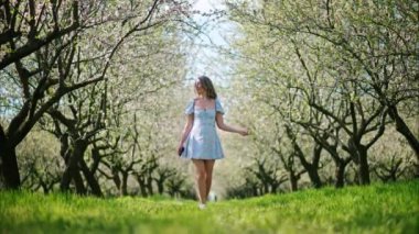 Mavi elbiseli esmer kadın çiçek açan badem ağaçlarının arasında yürüyor.