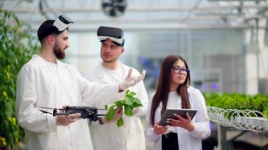 Beyaz önlüklü üç laboratuvar teknisyeni Sanal Gerçeklik kulaklığı takıyor, bir dron ve tablet tutuyor, serada yetişen marulu analiz ediyorlar.