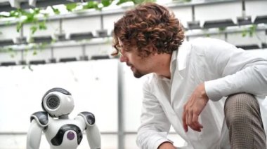 Beyaz önlüklü bir laboratuvar teknisyeni sera çiftliğindeki insansı robotla etkileşime geçiyor.