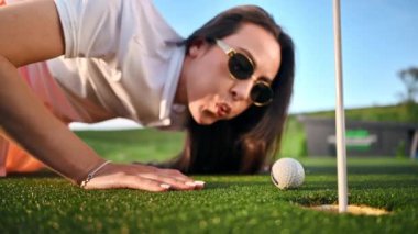 Bir kadın golf topunu üfleyerek deliğe sokmaya çalışıyor.