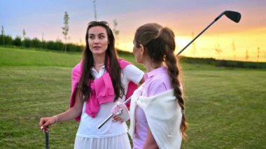 Beyaz ve pembe elbiseli iki kadın, ellerinde golf sopalarıyla sahada konuşuyorlar.