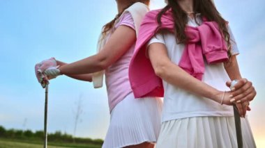 Beyaz ve pembe elbiseli iki kadın ellerinde golf sopalarıyla çimen tarlasında sırt sırta dururlar.