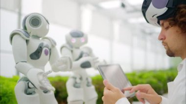 Beyaz önlüklü bir laboratuvar teknisyeni, sanal gerçeklik kulaklığı takıyor. Tablet üzerinde grafikleri analiz ederken bir sera çiftliğindeki iki insansı robotla iletişim kuruyor.