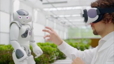 Beyaz önlüklü bir laboratuvar teknisyeni sanal gerçeklik kulaklığı takıyor ve bir seradaki farklı marul türlerinin yakınında insansı robotlarla etkileşime giriyor.