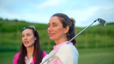 Beyaz ve pembe elbiseli iki kadın, ellerinde golf sopalarıyla sahada konuşuyorlar.