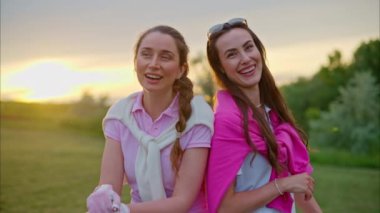 Beyaz ve pembe elbiseli iki kadın, ellerinde golf sopalarıyla sahada gülüyorlar.