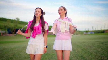 Beyaz ve pembe elbiseli iki kadın, ellerinde golf sopalarıyla sahada konuşuyorlar.