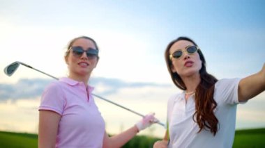 Beyaz ve pembe elbiseli iki kadın, ellerinde golf sopalarıyla sahada konuşuyorlar.