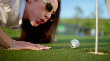 Bir kadın golf topunu üfleyerek deliğe sokmaya çalışıyor.