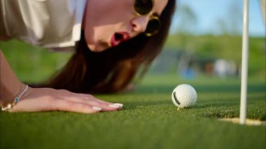 Bir kadın golf topunu üfleyerek deliğe sokmaya çalışıyor.