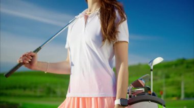 Beyaz ve pembe giyinmiş bir kadın omuzunda golf sopasıyla sahada dikiliyor.