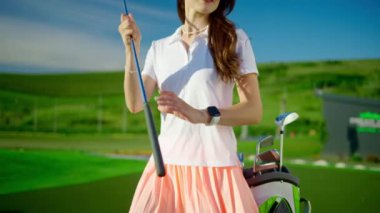 Beyaz ve pembe giyinmiş bir kadın golf sopasını golf sahasında bir çantaya koyuyor.
