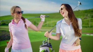Omuzlarında golf sopaları olan, beyaz ve pembe elbiseli iki kadın golf sahasında gülüyorlar.