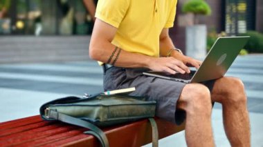 Sarı gömlekli bir adam bankta dikilip dizüstü bilgisayarla konuşuyor.