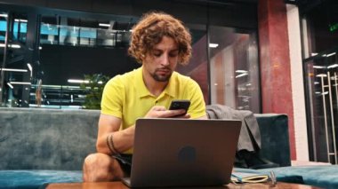 Sarı tişörtlü bir adam kanepeye oturmuş telefonuyla oynaşıyor ve bir dizüstü bilgisayar üzerinde çalışıyor.