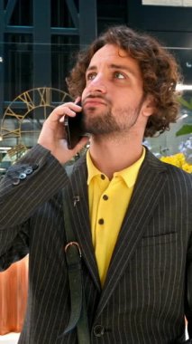 Sarı gömlekli ve gri ceketli bir adam ofis binasında telefonla konuşuyor. Dikey