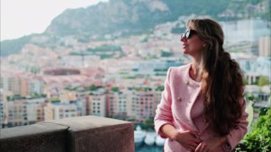 Pembe ceketli esmer kadın Monako 'daki Port de Fontvieille' e yanaşmış teknelerin bulanık görüntüsüyle gülümsüyor.