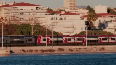 Antibes, Fransa - 14 Kasım 2024: Sahildeki binaların ve palmiye ağaçlarının önünde hareket eden tren arka plandaki dağları görüyor