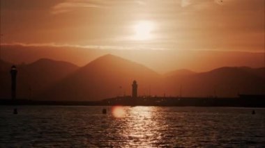 Gün batımında arka planda dağların olduğu uzak bir deniz feneri manzarası