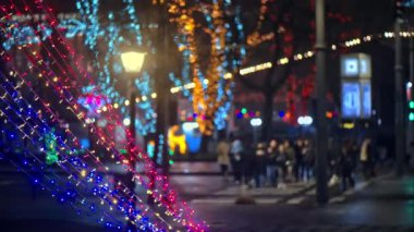 Arka planda karşıdan karşıya geçen insanların bulanık bir görüntüsüyle parıldayan renkli Noel ışıklarını kapat